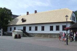 Budynek z białą elewacją i nowym jasnym dachem pokrytym gontem. Przed wejściem schody i daszek. W tle drzewa. Po prawej stronie grupka osób i latarnia uliczna. Na placu brukowana nawierzchnia.
