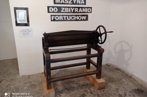 Zabytkowa maszyna z metalowym walcem i korbą, osadzona na drewnianej ramie. Ustawiona na kamiennej podłodze przy białej ścianie. Nad nią napis: „Maszyna do zbiyranio fortuchów”. Obok tabliczka informacyjna.