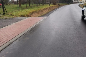 Droga asfaltowa po remoncie z przylegającym chodnikiem z kostki brukowej po lewej stronie. Po prawej stronie fragment pojazdu, dalej teren zielony i drzewa. W oddali linie energetyczne i zabudowa jednorodzinna. Nawierzchnia wilgotna, pogoda pochmurna.