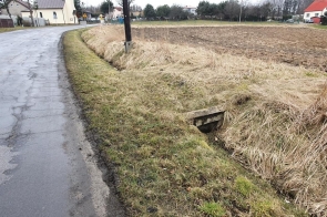 Zdjęcie przedstawia wiejską drogę asfaltową biegnącą obok pola uprawnego. Po prawej stronie widać rowy melioracyjne z betonowym przepustem. W tle znajdują się domy jednorodzinne oraz drzewa. Pogoda pochmurna, nawierzchnia wilgotna.