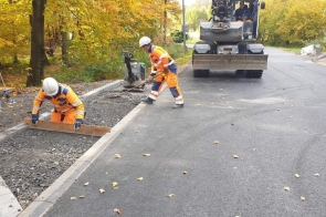 Prace wykończeniowe przy nowo wybudowanej drodze. Dwóch pracowników w odzieży ochronnej montuje elementy infrastruktury drogowej przy krawężniku. Widoczna świeżo ułożona nawierzchnia asfaltowa oraz sprzęt budowlany w tle. Otoczenie zalesione, liście w kolorach jesieni.
