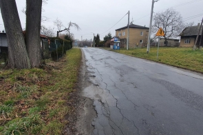 Droga asfaltowa przebiegająca przez teren zabudowany. Po lewej stronie drzewa i ogrodzenia posesji, po prawej budynki mieszkalne i gospodarcze. Widoczne znaki drogowe. Nawierzchnia wilgotna, niebo zachmurzone.