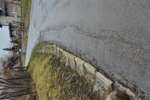 Droga asfaltowa z zakrętem, po prawej stronie uszkodzony krawężnik z betonowych elementów. Po lewej stronie drzewa bez liści oraz budynek mieszkalny. Nawierzchnia drogi wilgotna, niebo zachmurzone.