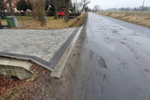 Droga asfaltowa z wjazdem z kostki brukowej prowadzącym do posesji po lewej stronie. Widoczny betonowy przepust pod zjazdem. Po prawej stronie pola uprawne. Nawierzchnia drogi wilgotna, niebo zachmurzone.