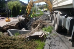 Prace ziemne przy drodze z użyciem koparki i samochodu ciężarowego. Widoczne elementy betonowe przygotowane do montażu. Po lewej stronie posesje, po prawej pas zieleni. Teren wykopu częściowo uprzątnięty, robotnicy w trakcie pracy.