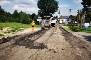 Odcinek drogi w trakcie remontu. Na jezdni widoczna zrywka starej nawierzchni asfaltowej. Prace prowadzone z użyciem maszyny drogowej i obecnością pracownika w odzieży ochronnej. W tle zabudowa miejscowości i znaki drogowe.