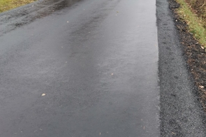 Droga asfaltowa po zakończonym remoncie. Nawierzchnia równa i ciemna, świeżo ułożona. Po obu stronach drogi pobocza gruntowe z roślinnością. W tle widoczność ograniczona przez mgłę. Otoczenie wiejskie z pojedynczymi drzewami.