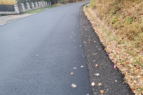 Droga asfaltowa po zakończeniu prac remontowych. Nawierzchnia równa, bez uszkodzeń. Po prawej stronie skarpa porośnięta trawą i drzewami, po lewej barierki ochronne. Na jezdni widoczne pojedyncze liście. Otoczenie leśne, jesienna szata roślinności.