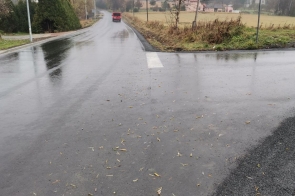 Skrzyżowanie dróg asfaltowych po zakończeniu prac remontowych. Nawierzchnia równa i wilgotna. Po lewej stronie lampa uliczna i posesja, po prawej pole uprawne oraz znak drogowy z nazwą ulicy. Teren otwarty, widoczność dobra mimo pochmurnej pogody.