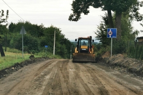 Droga w trakcie remontu, nawierzchnia przygotowana do dalszych prac. Na środku jezdni spycharka. Po obu stronach drogi widoczne znaki drogowe oraz zieleń. Brak ruchu pojazdów, teren budowy.