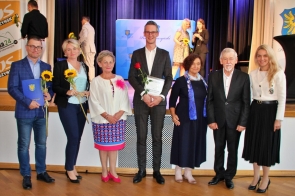Grupa siedmiu osób pozuje do zdjęcia podczas oficjalnej uroczystości. Trzy osoby trzymają kwiaty oraz dyplomy, a w tle widać elementy dekoracyjne oraz logo Powiatu Cieszyńskiego.