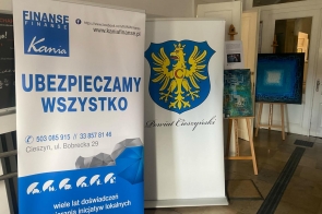 Zdjęcie banerów, od lewej baner firmy Kania Finanse, następnie Powiat Cieszyński.  Na drugim planie stoi obraz na drewnianej sztaludze.  