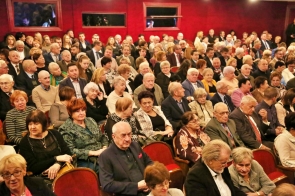 Duża grupa osób siedzi w rzędach na czerwonych fotelach w sali widowiskowej. Większość jest zwrócona w stronę sceny, część rozmawia między sobą. Wśród zgromadzonych widoczne są osoby w różnym wieku, głównie w eleganckich strojach.