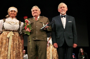 Trzy osoby stoją obok siebie na scenie. Pośrodku mężczyzna w mundurze trzyma różę oraz otwarte etui z wyróżnieniem, po lewej stoi kobieta w stroju regionalnym, a po prawej mężczyzna w garniturze z muszką. W tle widoczne są inne osoby uczestniczące w uroczystości.