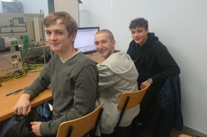 Trzech uczniów siedzi przy stanowisku wyposażonym w komputer oraz urządzenia elektroniczne z licznymi przewodami. Wszyscy są zwróceni w stronę aparatu, a w tle widoczny jest monitor i panel laboratoryjny.