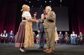 Kobieta w stroju regionalnym podaje rękę mężczyźnie w mundurze, który trzyma ozdobne etui. W tle na scenie siedzą inne osoby, część z nich trzyma kwiaty. Całość odbywa się na tle czerwonych kurtyn i dużego ekranu.