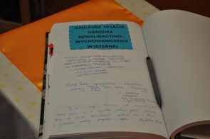 Otwarta księga pamiątkowa leży na stole. Na lewej stronie widoczne są odręczne wpisy oraz niebieska kartka z napisem „Jubileusz 10-lecia Ośrodka Rewalidacyjno-Wychowawczego w Istebnej”. Obok leży długopis.