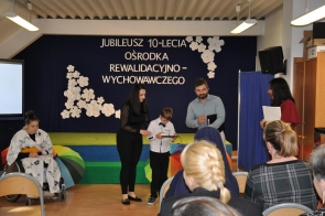Chłopiec stoi przed sceną i trzyma dokument, obok niego stoją kobieta w czarnym stroju oraz mężczyzna z teczką. Po lewej stronie siedzi osoba na wózku. W tle widoczna jest granatowa dekoracja z napisem i białymi kwiatami.