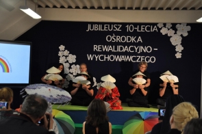 Grupa osób występuje na scenie podczas jubileuszu 10-lecia Ośrodka Rewalidacyjno-Wychowawczego. Uczestnicy mają wachlarze i stożkowe nakrycia głowy, a jedna osoba siedzi pośrodku w czerwonym stroju. Przed sceną widoczne są kolorowe parasole, a w tle granatowa dekoracja z białym napisem i kwiatami.
