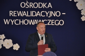 Mężczyzna w garniturze przemawia do mikrofonu, trzymając czerwoną teczkę. W tle widoczna jest granatowa dekoracja z dużym napisem „Ośrodka Rewalidacyjno-Wychowawczego” oraz białymi kwiatami.