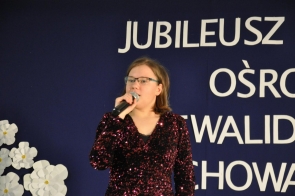 Kobieta w okularach śpiewa do mikrofonu. Ma na sobie ciemną, połyskującą bluzkę. W tle widoczna jest granatowa dekoracja z dużym białym napisem jubileuszowym i białymi kwiatami.