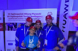 Cztery osoby stoją przed ścianką informacyjną Olimpiad Specjalnych. Trzy osoby mają niebieskie bluzy i czerwone czapki, a stojąca z przodu kobieta w niebieskiej koszulce pokazuje kciuki uniesione do góry.