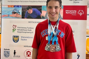 Mężczyzna w czerwonej koszulce stoi przed ścianką Olimpiad Specjalnych. Na szyi ma zawieszone liczne medale. W tle widoczne są zdjęcia związane z pływaniem oraz informacje o IX Ogólnopolskim Mityngu Pływackim i VII Ogólnopolskim Turnieju Bowlingowym.
