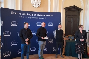 Cztery osoby stoją na tle granatowego banera z białym napisem o szkole i emblematami, w eleganckiej sali z jasnymi ścianami i godłem Polski. Dwóch mężczyzn w ciemnych ubraniach, jeden trzyma kartkę, a obok nich kobieta w sukience (dyr. Regina Rakowska) i mężczyzna w kamizelce (Michał Rajwa, Naczelnik Wydziału Edukacji). Na stole puchary.