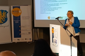 Na zdjęciu widoczna jest Anna Stefaniak-Bacza, dyrektor PUP w Cieszynie w niebieskim żakiecie, przemawiająca na tle ekranu z wyświetloną prezentacją pt. „Punkt dla młodzieży – Mentorzy”. W tle znajdują się banery promocyjne i logo. Przed kobietą siedzą słuchacze, z których część jest widoczna od tyłu.