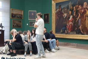 Grupa młodych ludzi siedzi na ławce w galerii sztuki w Krakowie, otoczona obrazami o treści historycznej, zawieszonymi na zielonych ścianach. Pomieszczenie jest przestronne, dobrze oświetlone, dzięki dużym oknom. W dolnym rogu znajduje się logo „Podróże z klasą”, co oznacza, że uczestnicy wycieczki są uczestnikami programu „Podróże z Klasą”.
