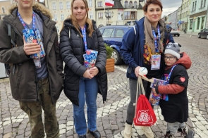 Grupa czterech osób stoi na miejskim placu, trzymając kolorowe puszki Wielkiej Orkiestry Świątecznej Pomocy. Uśmiechnięte twarze i odznaki wolontariuszy dodają energii zdjęciu. Wśród nich młody chłopiec w czapce i kurtce z czerwonym plecakiem. W tle widać budynki z flagami i brukowaną nawierzchnię placu, co podkreśla zimową aurę i atmosferę zaangażowania.