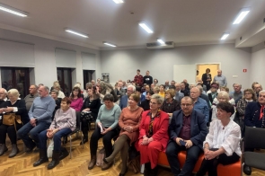 Sala pełna osób siedzących na krzesłach, skierowanych w stronę prelegenta, którego nie widać na zdjęciu. Grupa obejmuje osoby w różnym wieku, głównie starsze, ubrane w codzienne lub eleganckie stroje. W tle widoczne są białe ściany, okna z roletami oraz otwarte drzwi, przez które widać stojące w grupie osoby. Pomieszczenie jest jasno oświetlone.