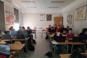 Uczniowie siedzą w jasnej klasie przy ustawionych w rzędach ławkach, pracując na laptopach. Na ścianach wiszą mapy i plakaty edukacyjne. Część uczniów w bluzach, skupieni na ekranach. Na podłodze leżą plecaki i podłączone do komputerów kable zasilające.