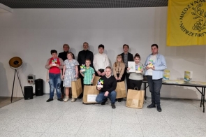 Grupa osób pozuje do zdjęcia we wnętrzu sali z białą ścianą i żółtym banerem. Część uczestników trzyma kolorowe układanki w kształcie puzzli oraz torby z upominkami. W tle stolik z nagrodami i kilkoro dorosłych opiekunów.