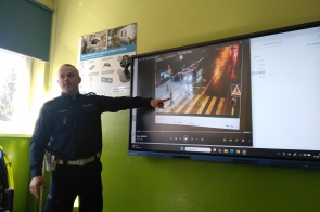 Policjant w ciemnym mundurze prowadzi prelekcję w klasie, wskazując na ekranie moment potrącenia pieszego na pasach. W tle zielona ściana, plakat techniczny i światło dzienne wpadające przez okno. Pokaz dotyczy bezpieczeństwa na drodze.
