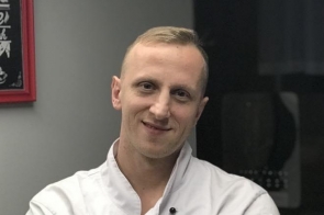 Łukasz w białym fartuchu kucharskim z założonymi rękami stoi na tle szarej ściany. Uśmiechnięty, z krótkimi blond włosami i zegarkiem na żółtym pasku prezentuje się pewnie i profesjonalnie.