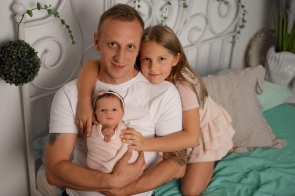 Łukasz w białej koszulce siedzi na łóżku, trzymając na rękach noworodka owiniętego w jasny kocyk. Obok niego przytula się kilkuletnia dziewczynka w jasnoróżowej sukience, obejmując tatę i młodszą siostrę z czułością i uśmiechem.