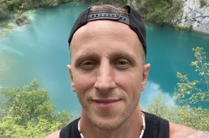 Uśmiechnięty Łukasz w czapce z daszkiem do tyłu robi selfie na tle turkusowego jeziora otoczonego zielenią i wapiennymi skałami. Ma na sobie czarną koszulkę bez rękawów i biały naszyjnik z koralików, a na ramieniu widoczny tatuaż.