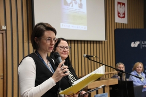Od lewej Beata Szymańska, główna specjalistka ds. promocji zdrowia oraz Olga Białoń, prezeska Stowarzyszenia „Pomocna Dłoń” - stoją przy mównicy, trzymając mikrofony i dokumenty podczas konferencji w sali Uniwersytetu Śląskiego. W tle widać slajd prezentacji oraz godło Polski. Obok mównicy siedzą dwie inne prelegentki przy stole z komputerem. 