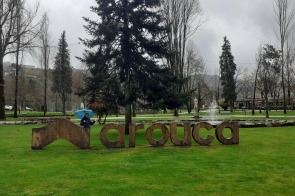 Zielony park z dużym napisem „Arouca” z drewnianych liter ustawionych na trawie. W tle wysokie drzewa bez liści, alejki i fontanna. Po lewej stronie osoba z niebieskim parasolem pozuje przy napisie, mimo pochmurnej i deszczowej pogody.