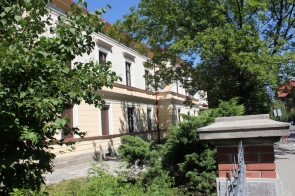 Budynek z jasną, świeżo odnowioną elewacją, otoczony zielenią; widoczna część ogrodzenia z cegły i metalu oraz fragment chodnika. Zdjęcie po zakończeniu termomodernizacji.