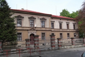 Dwukondygnacyjny, zabytkowy budynek z czerwonym dachem, elewacją w odcieniach beżu i brązu, ozdobnymi gzymsami nad oknami oraz głównym wejściem pośrodku. Przed budynkiem znajduje się ogrodzenie z metalową bramą i ceglanymi słupkami. Na pierwszym piętrze widać firanki w oknach, a na elewacji zamontowana jest antena satelitarna. Po bokach rosną drzewa, a ulica biegnie wzdłuż ogrodzenia. Zdjęcie przed termomodernizacją.