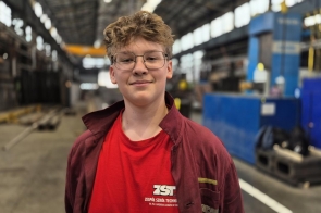 Uczeń ubrany w czerwoną koszulkę i uniform szkoły technicznej, z uśmiechem stoi w przestronnej hali fabrycznej. W tle widoczne są maszyny i konstrukcje przemysłowe. Jest zadowolony z warunków w trzynieckim zakładzie.