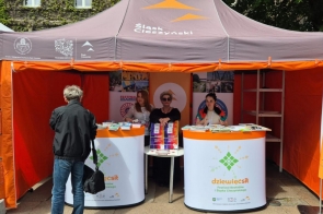 Stół pokryty stosami kolorowych broszur i ulotek, promujących atrakcje turystyczne. W tle plakat z logo festiwalu rowerowego. Dominują jasne i żywe kolory, tworząc przyciągającą wzrok ekspozycję na pomarańczowym tle.