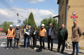 Grupa osób stoi przed budynkiem oznaczonym jako Przedszkole nr 7 w Cieszynie. Wśród nich są osoby w odzieży roboczej i kamizelkach odblaskowych. Tło stanowi zielona okolica i fragment drogi z oznaczeniami. Atmosfera jest formalna i słoneczna.