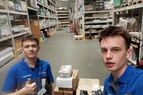 Dwaj młodzi mężczyźni, ubrani w niebiesko-szare koszulki z napisem „UCZEŃ”, stoją w przestronnej hali magazynowej między wysokimi regałami. Franek robi selfie, a Mateusz pokazuje gest uniesionego kciuka. Przed nimi ułożone są kartony.