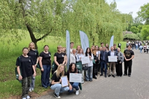 rupa młodzieży i nauczycieli pozuje na tle zielonych drzew z dyplomami i tablicami informującymi o nagrodzie w konkursie „Zielona Pracownia”. Uczestnicy są uśmiechnięci, w tle widoczni są spacerujący ludzie i drewniana chata.