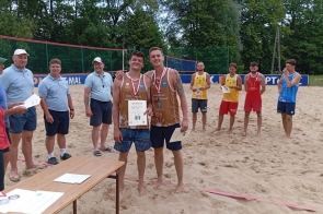 Dwóch młodych siatkarzy w brązowych koszulkach pozuje z medalami i dyplomem, obejmując się z uśmiechem. Obok nich organizatorzy, w tle inni zawodnicy czekający z dyplomami. Scena na piasku, w otoczeniu zielonych drzew i siatki.