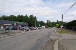 Widok na skrzyżowanie przy sklepie Delikatesy, z zatoczką autobusową po lewej i przejściem dla pieszych oznaczonym znakiem poziomym i pionowym. Ulica nosi ślady zużycia nawierzchni, widoczne są auta, piesi i słupy z kablami.