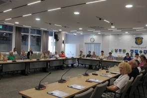 Uczestnicy obrad siedzą przy dużym, podkowiastego kształtu stole w sali konferencyjnej. Większość to seniorki, ubrane elegancko. Na ścianie herby gmin powiatu cieszyńskiego i zegar. Wnętrze jasne, nowoczesne i przestronne.
